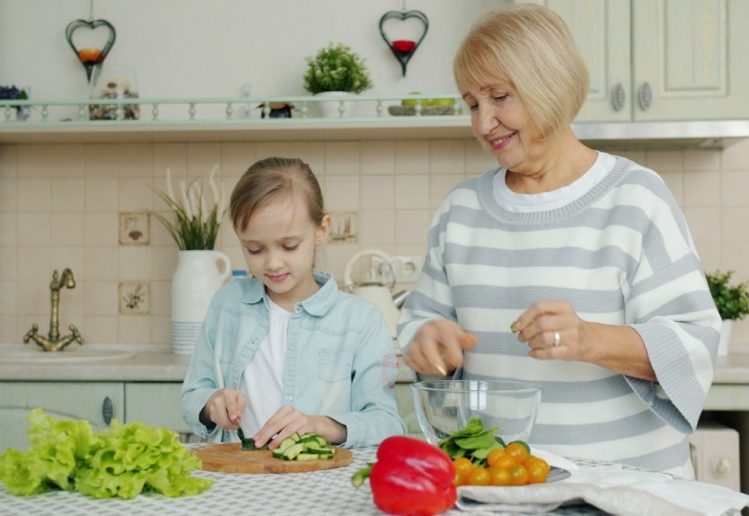 Granddaughter and grandmother are making salad together and talking in kitchen at home, little girl is cutting vegetables while old woman is watching her.
