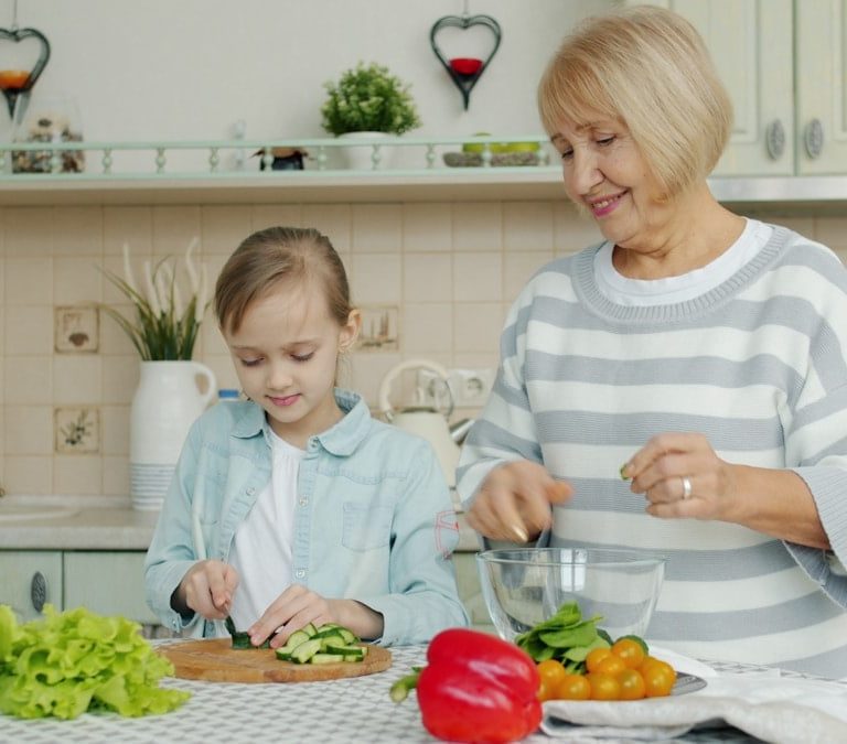 Granddaughter and grandmother are making salad together and talking in kitchen at home, little girl is cutting vegetables while old woman is watching her.