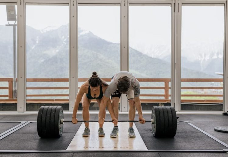 A couple lifting a heavy barbell together in a gym with mountain views.