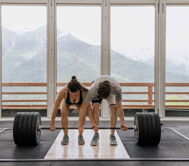 A couple lifting a heavy barbell together in a gym with mountain views.